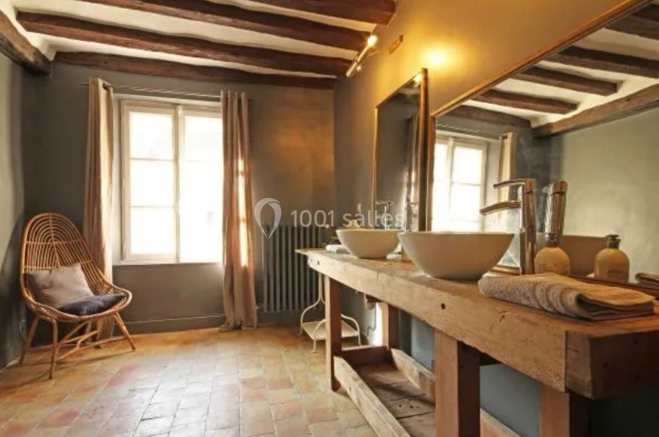 Salle de bain rustique avec double vasque sur meuble en bois, miroir large, chaise en rotin et lumière naturelle.