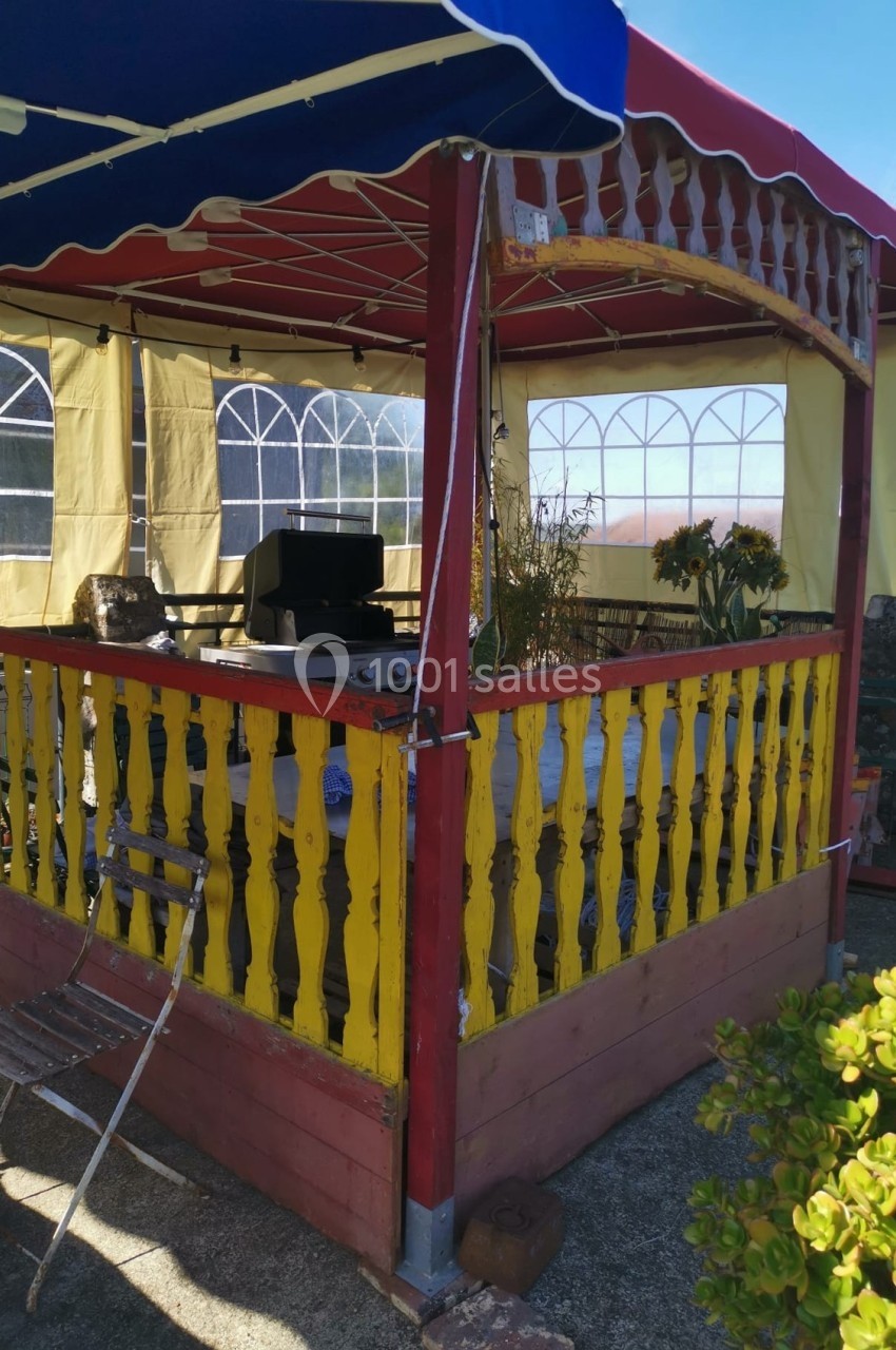 Tonnelle colorée avec balustrades en bois, abritant un barbecue et des plantes, située sur une terrasse ensoleillée.