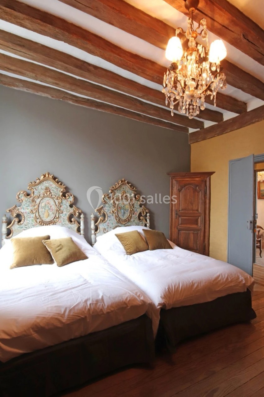 Chambre avec deux lits simples, têtes de lit décorées, poutres apparentes, armoire en bois et lustre suspendu.