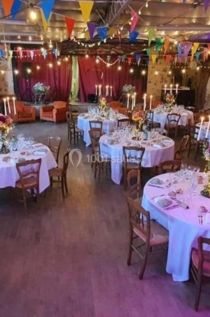 Salle décorée pour un événement, avec tables rondes dressées, chandeliers, guirlandes lumineuses et fanions colorés.