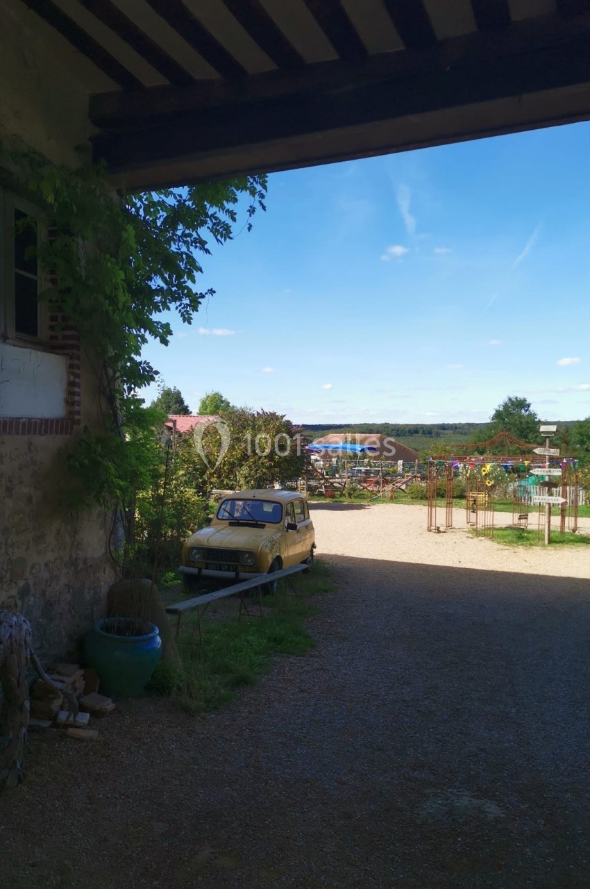 Vue d'une cour ensoleillée avec une vieille voiture jaune, des plantes et un paysage rural en arrière-plan.
