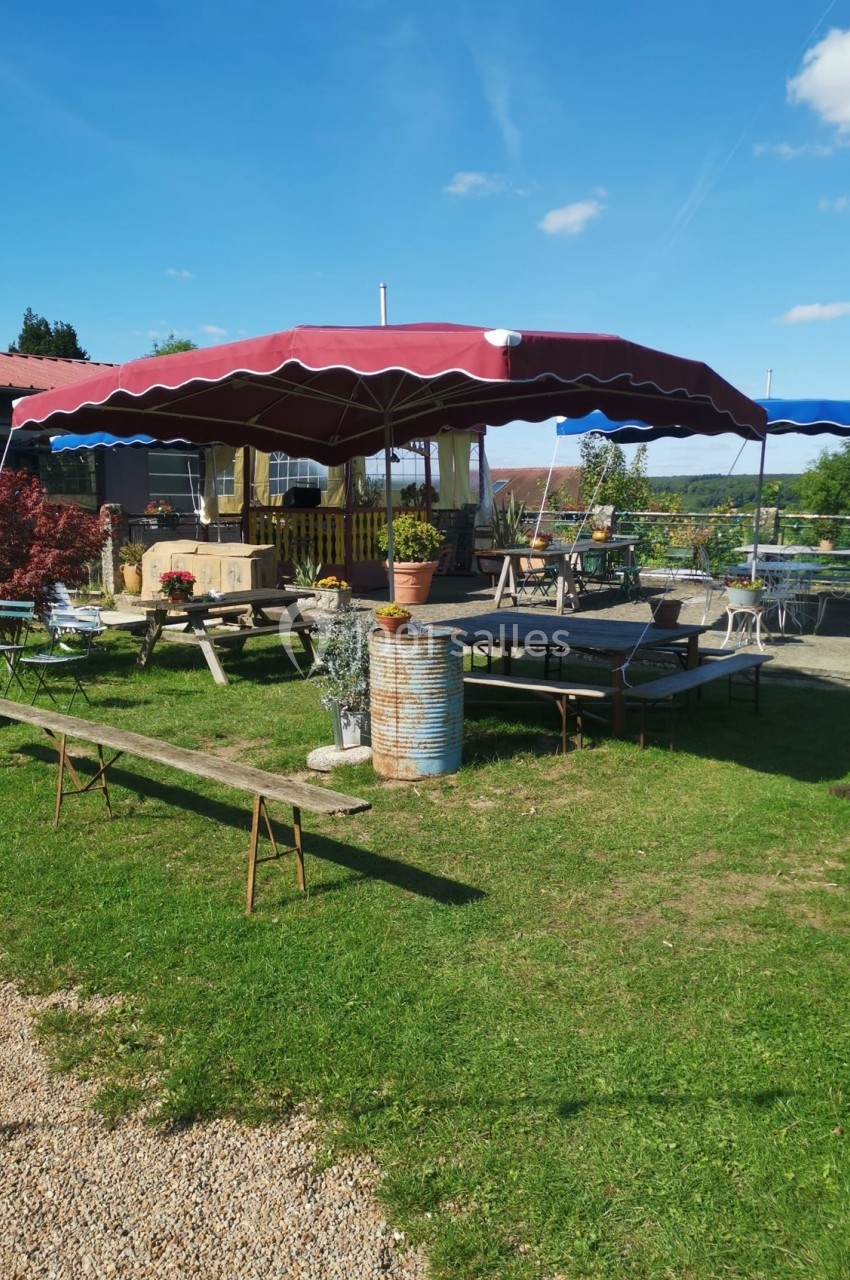 Terrasse extérieure avec tables en bois, bancs et parasols rouges, entourée de verdure sous un ciel bleu.