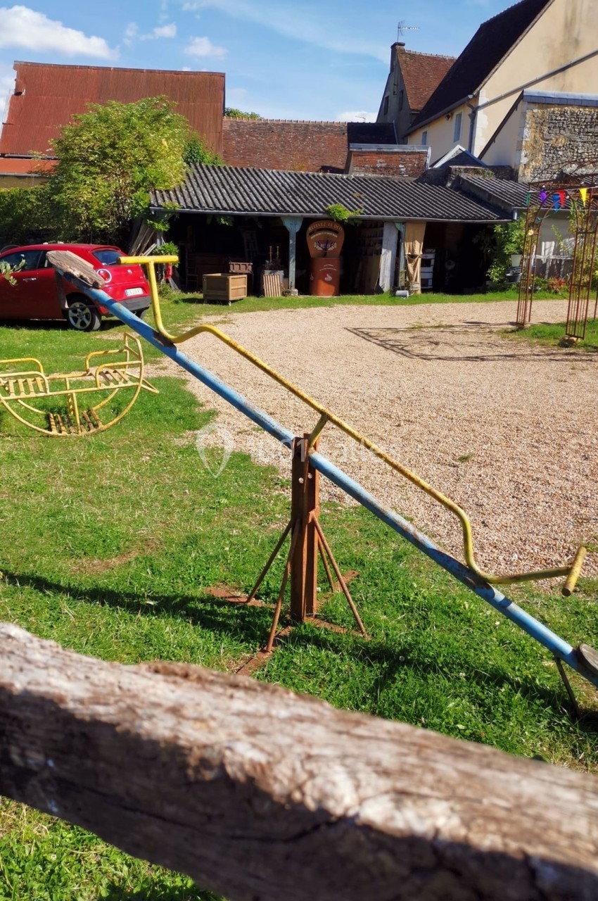 Balançoire à bascule en métal dans un jardin avec pelouse, graviers, bâtiments et voiture rouge en arrière-plan.