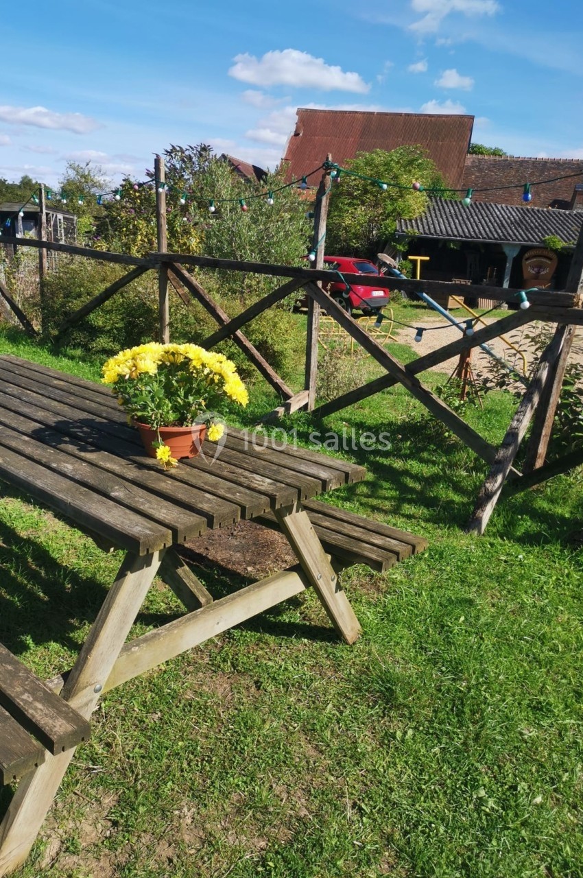 Table de pique-nique en bois avec un pot de fleurs jaunes, située dans un jardin ensoleillé avec des bâtiments en arrière…