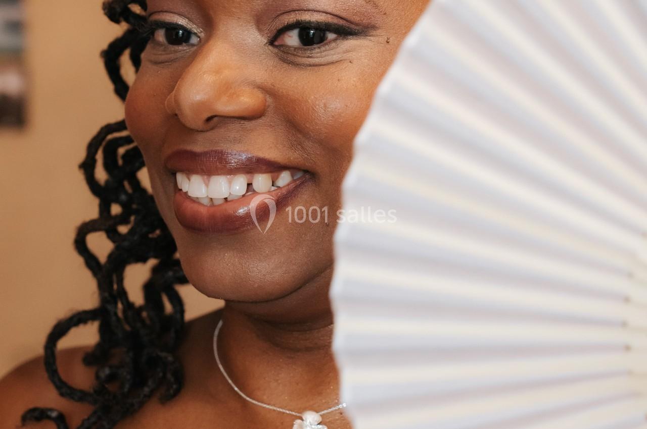 Femme souriante en robe blanche, portant un collier et une barrette ornée, tenant un éventail blanc devant elle.