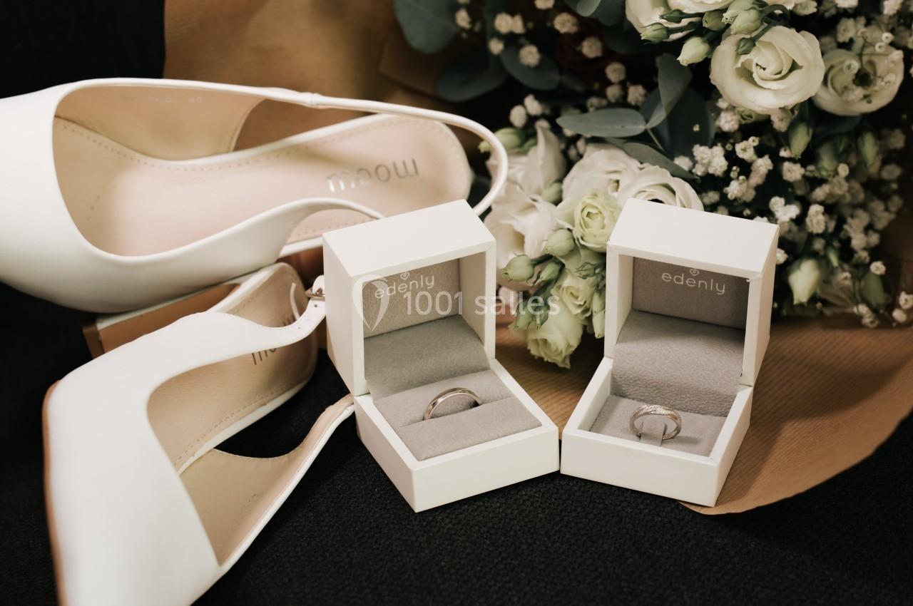 Deux alliances dans des écrins ouverts, entourées de chaussures blanches et d'un bouquet de fleurs.