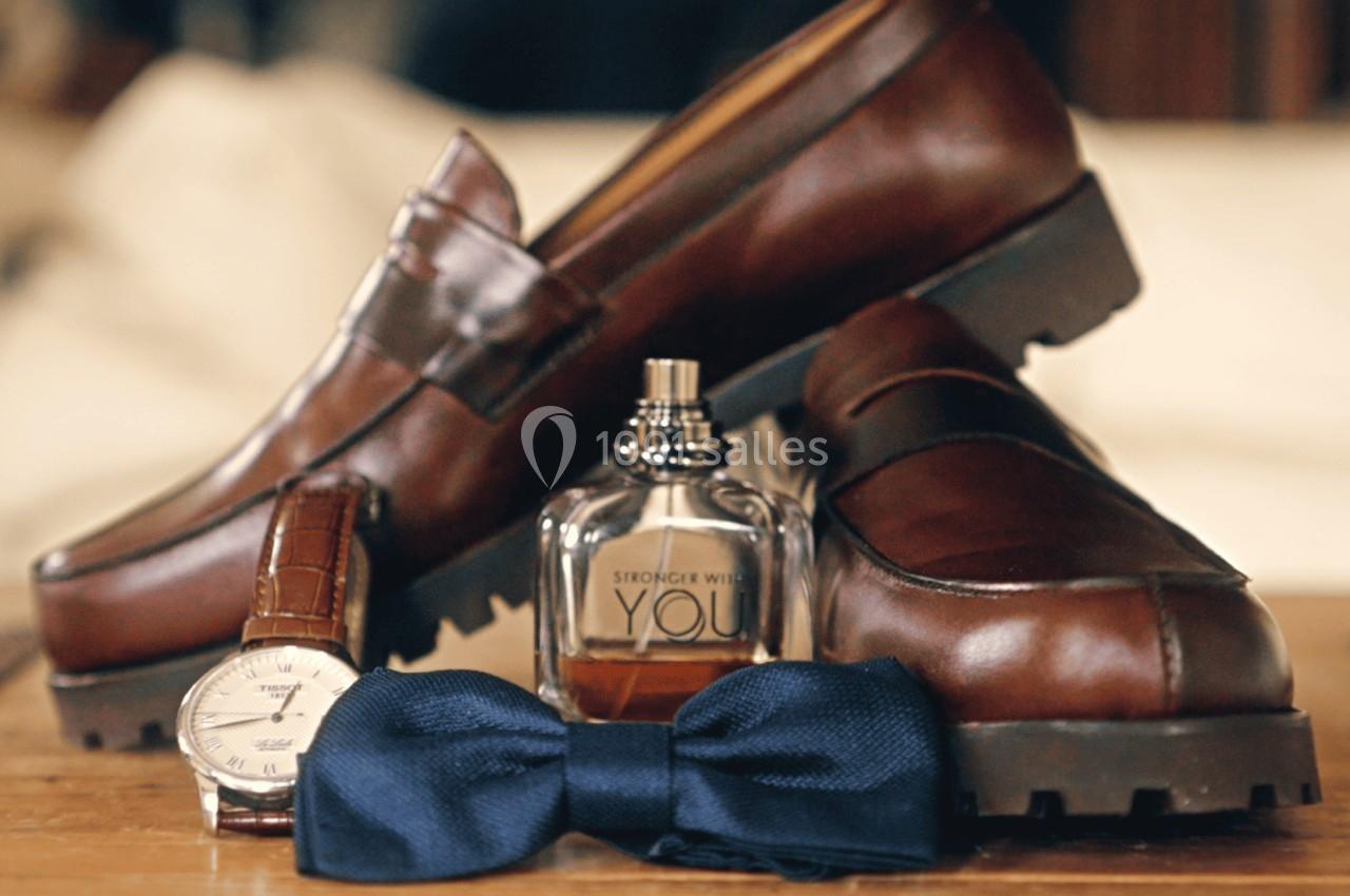 Chaussures en cuir marron avec une montre, un nœud papillon bleu et un flacon de parfum posés sur une table en bois.
