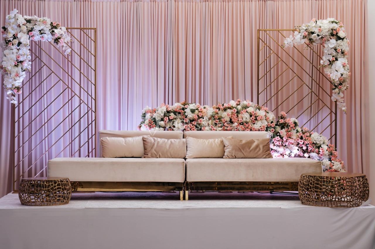 Canapé beige devant un rideau rose, décoré de fleurs blanches et roses, encadré par des structures dorées.