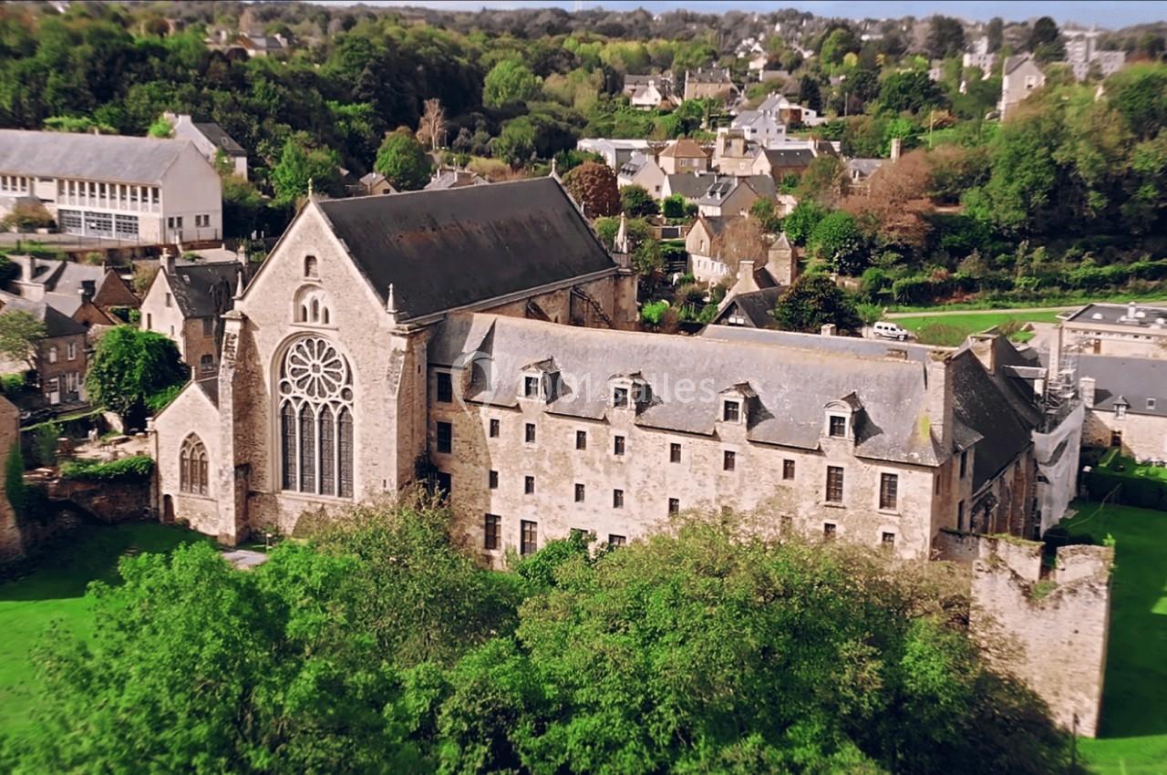 Vue aérienne d'une abbaye en pierre entourée de bâtiments historiques et de verdure dans un village.