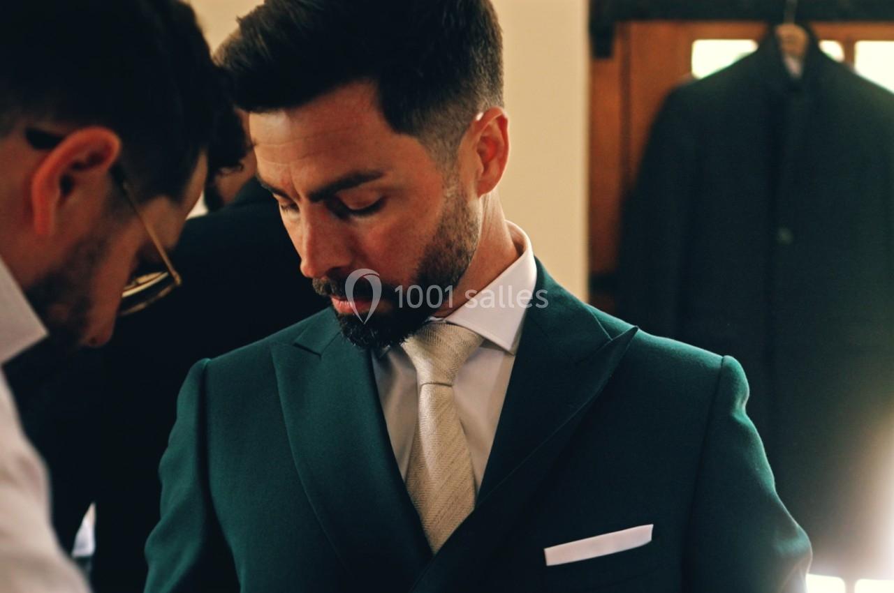 Un homme en costume vert est ajusté par une autre personne dans une pièce avec des vêtements suspendus en arrière-plan.