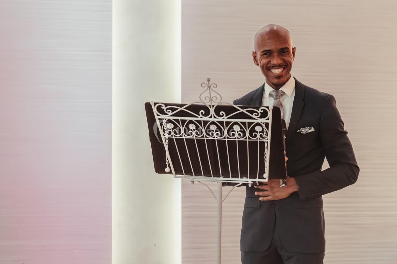 Homme souriant en costume sombre debout près d'un pupitre décoratif dans un intérieur lumineux.