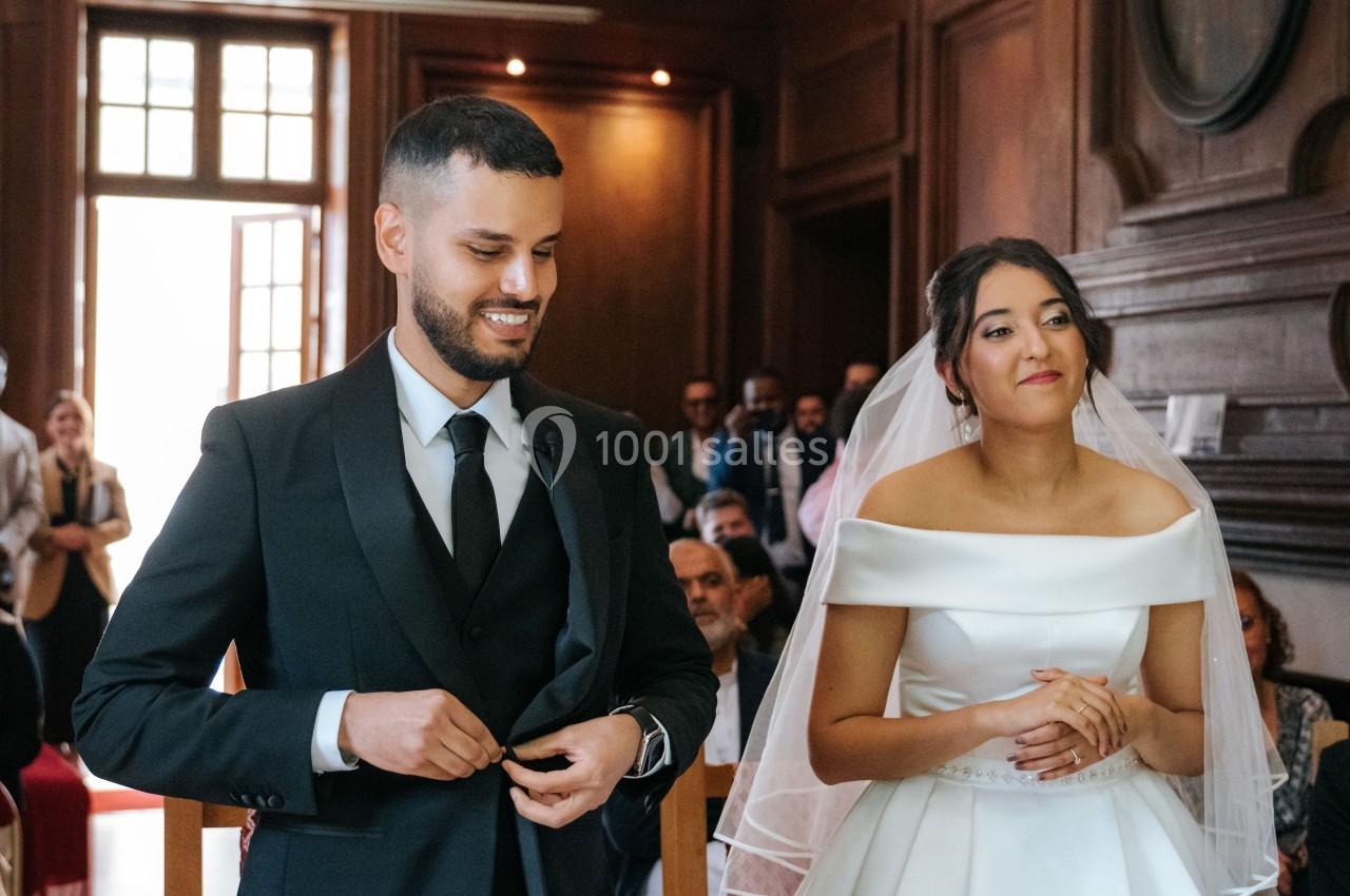 Un couple en tenue de mariage se tient debout dans une salle en bois, entouré de quelques invités.