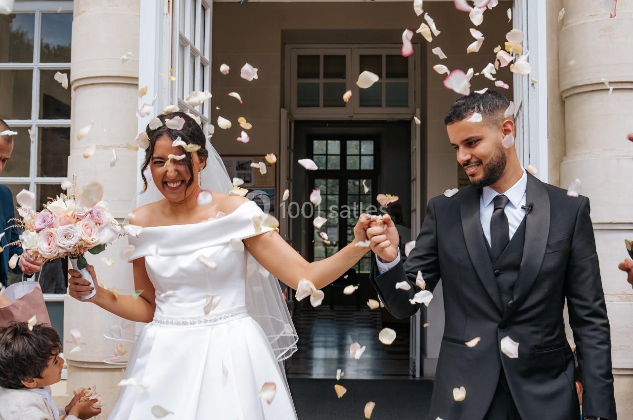 Un couple de mariés souriants sort d'un bâtiment sous une pluie de pétales de fleurs.