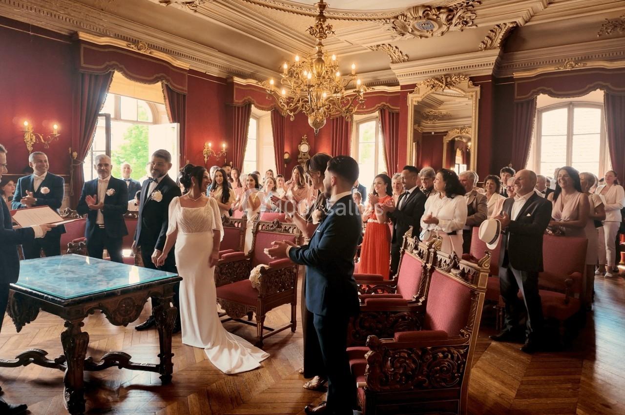 Un couple se marie dans une salle ornée avec des invités applaudissant, sous un éclairage naturel et des lustres élégants.