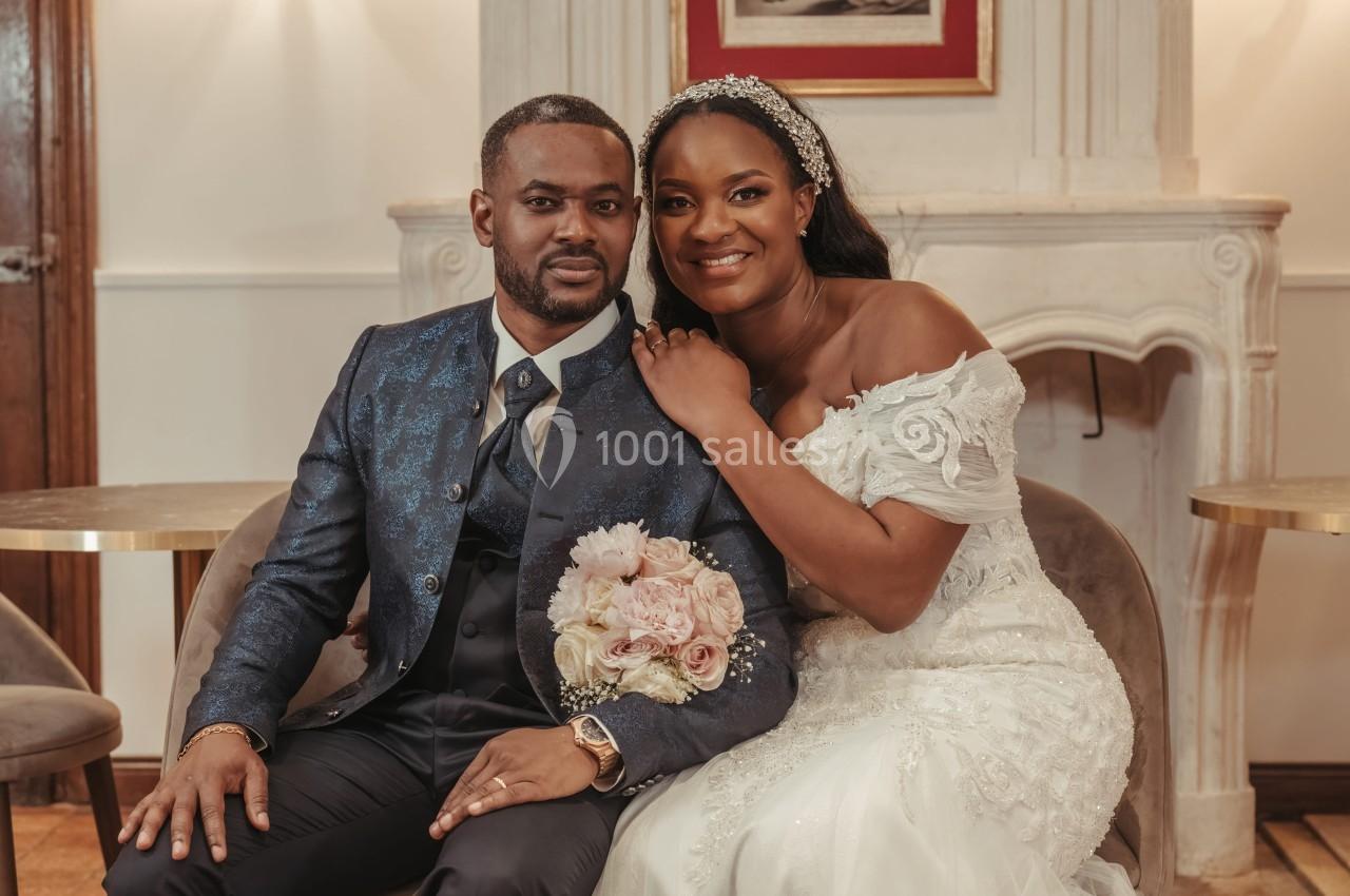 Un couple de mariés assis côte à côte, souriant, dans un décor intérieur élégant avec une cheminée en arrière-plan.