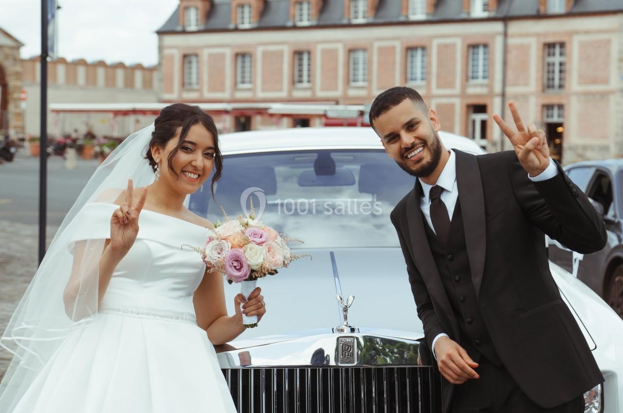 Un couple souriant en tenue de mariage pose devant une voiture blanche ornée d'un bouquet de fleurs.