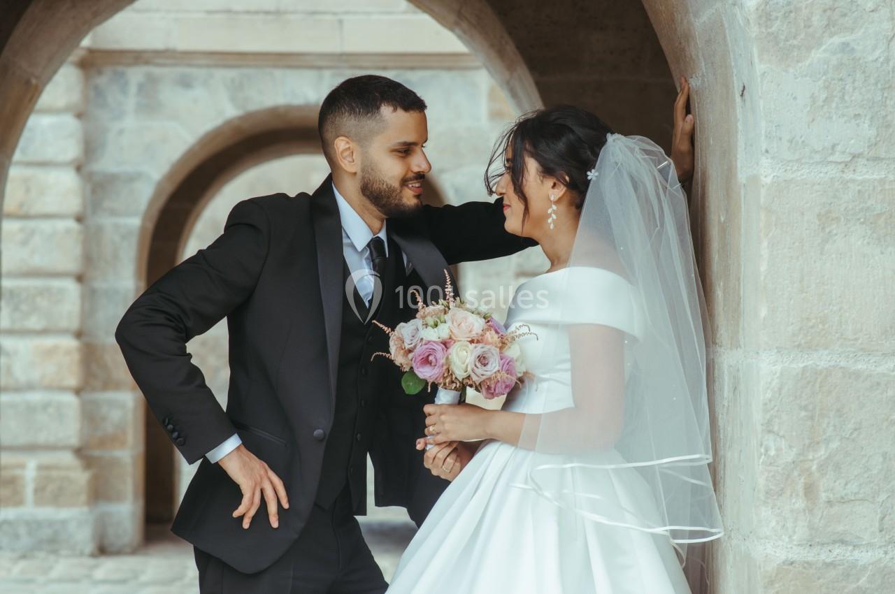 Un couple de mariés sourit et échange un regard complice sous une arche en pierre, la mariée tenant un bouquet.