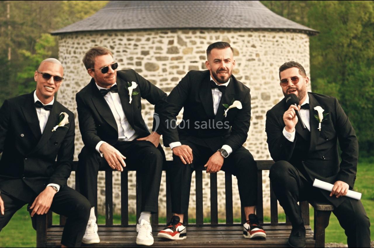 Quatre hommes en costume noir assis sur un banc devant un bâtiment en pierre, dans un cadre verdoyant.