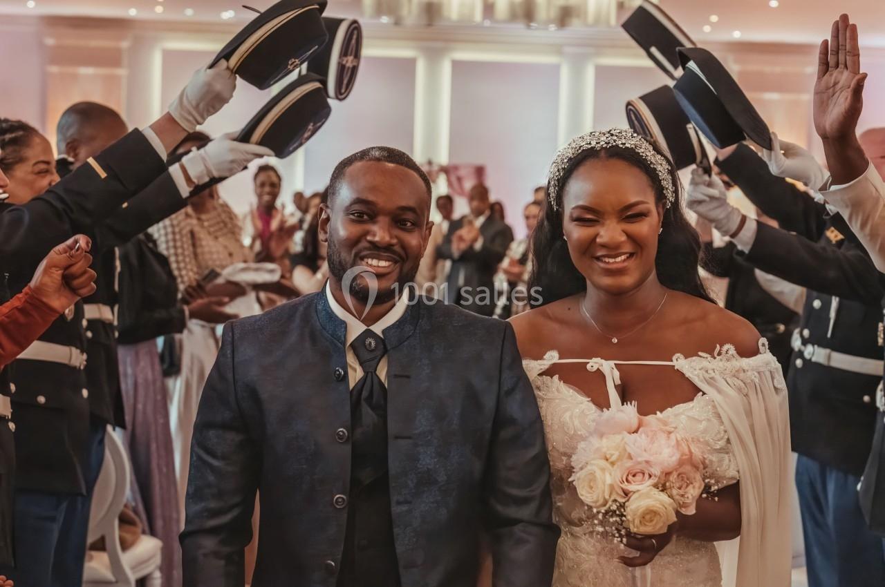 Un couple souriant en tenue de mariage avance sous une haie d'honneur formée par des militaires levant leurs képis.