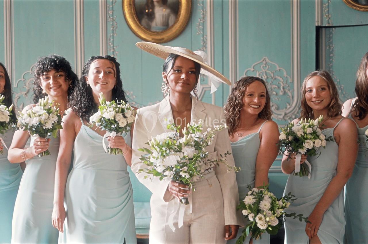 Un groupe de femmes en robes pastel et une femme en costume blanc posent avec des bouquets dans une salle élégante.
