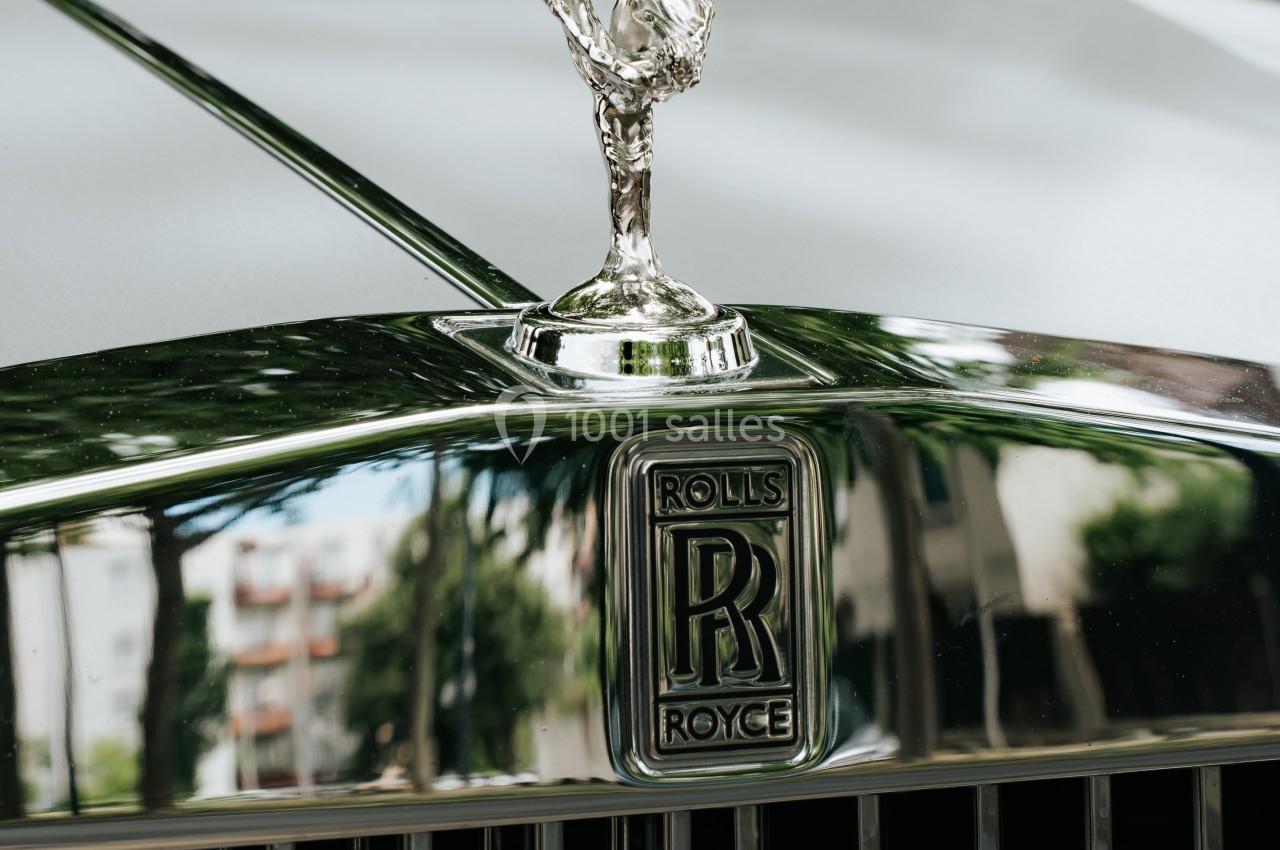 Ornement argenté ’Spirit of Ecstasy’ sur le capot d'une voiture Rolls-Royce, avec des reflets d'arbres et de bâtiments.