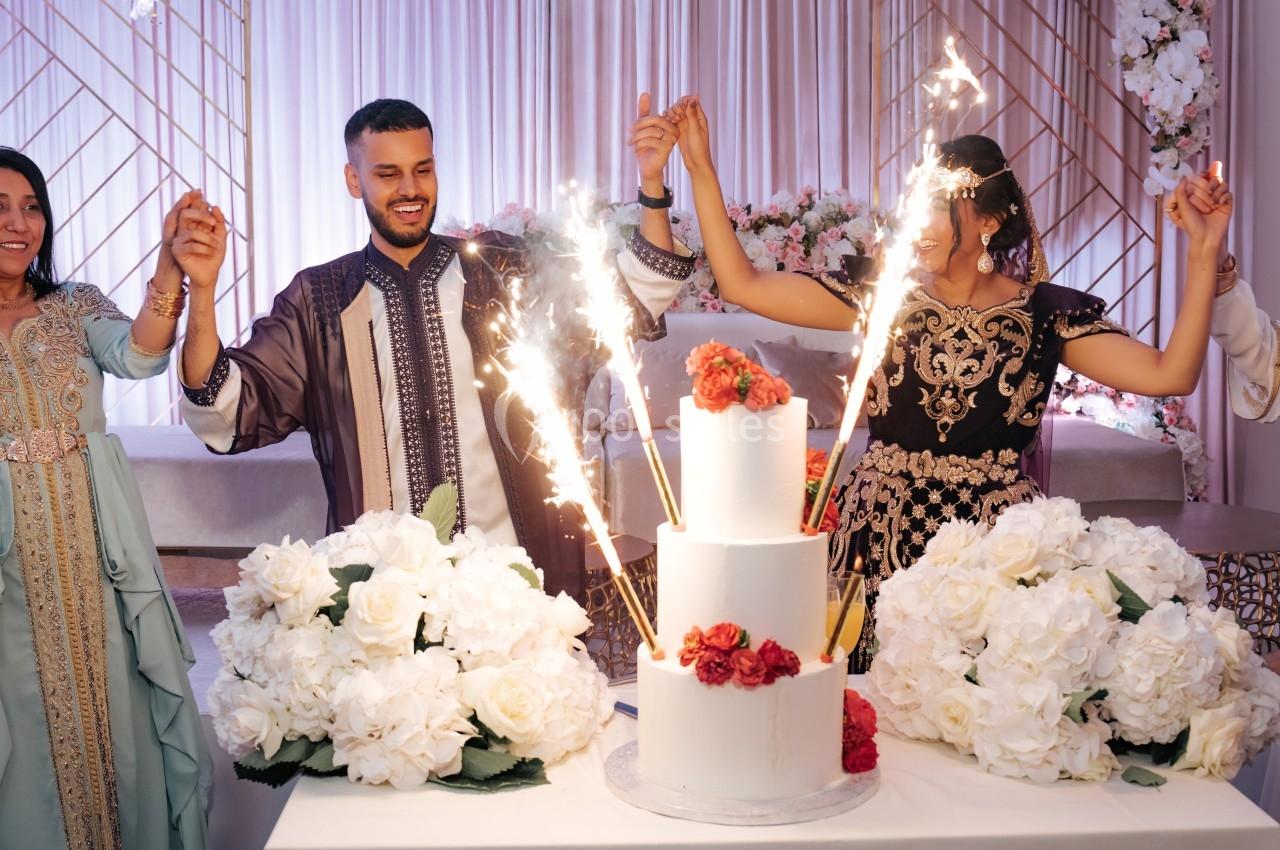 Un couple en tenue traditionnelle célèbre devant un gâteau décoré de fleurs et de fontaines lumineuses.
