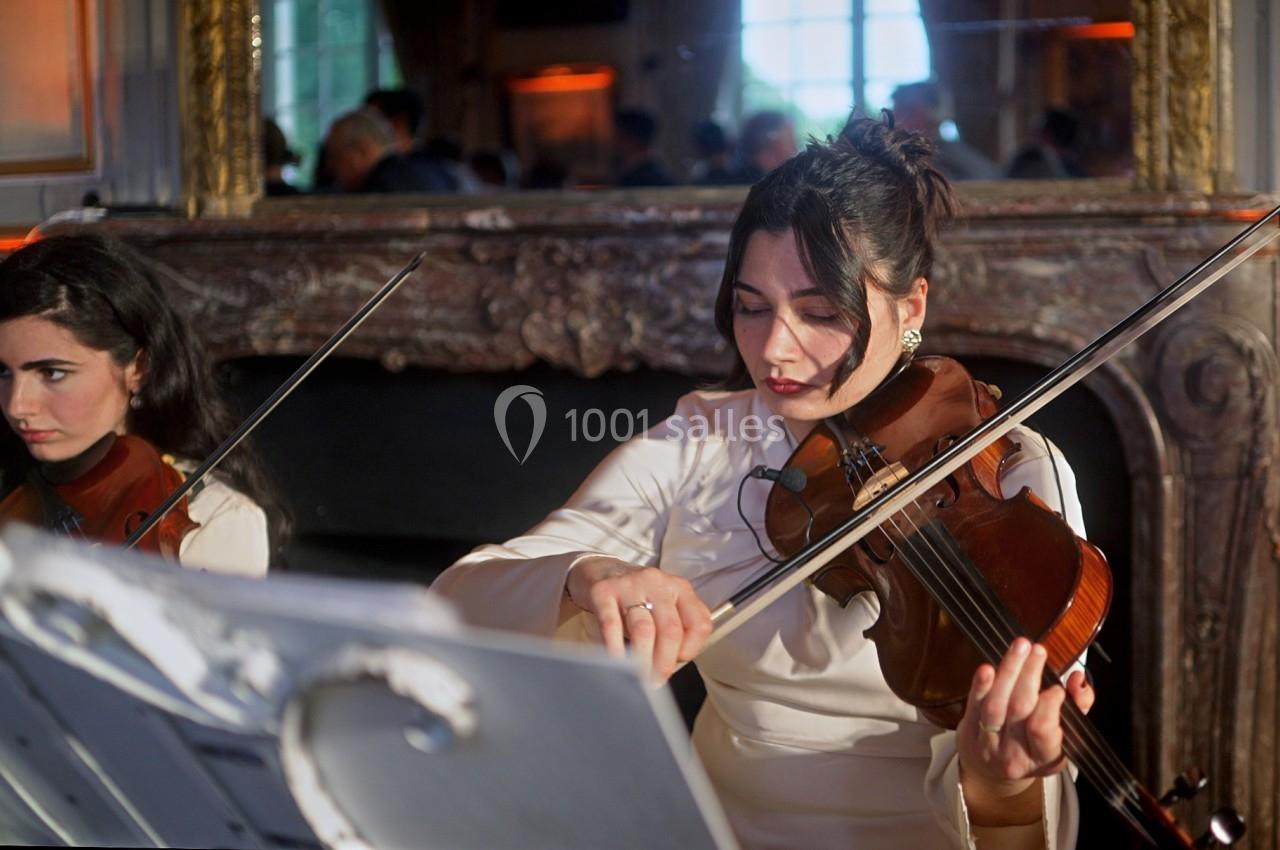 Deux musiciennes jouent du violon dans une pièce élégante avec un miroir et une cheminée en arrière-plan.