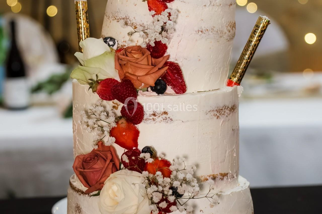 Gâteau de mariage à trois étages décoré de fleurs, fruits rouges et bâtonnets dorés, posé sur une table noire.