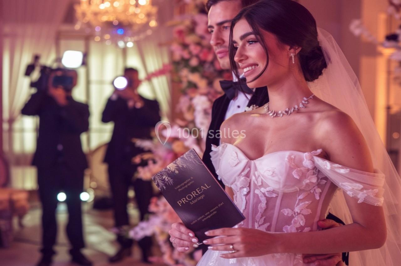 Une mariée souriante en robe blanche tient un carton, posant avec un homme en costume dans un décor de mariage éclairé.