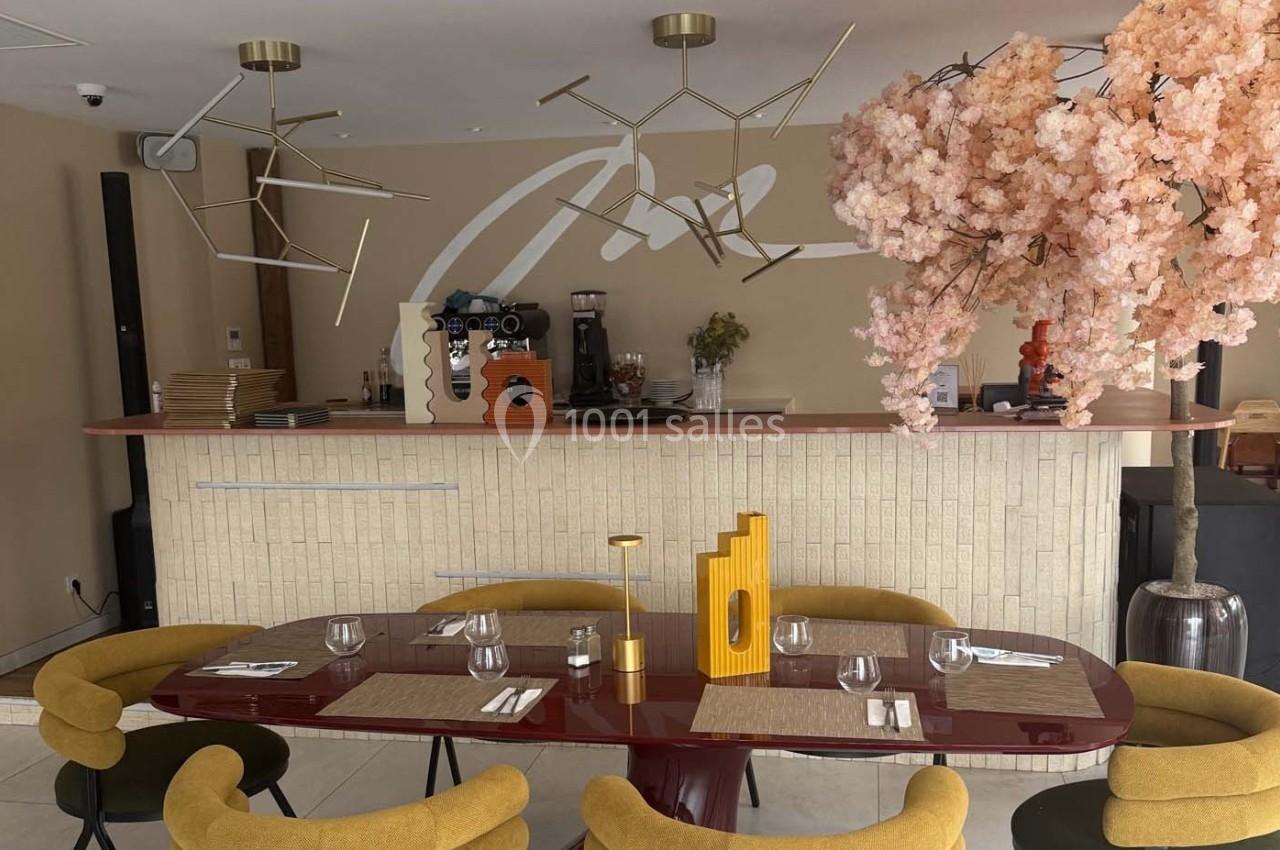 Salle de restaurant moderne avec comptoir en arrière-plan, table dressée et décoration florale rose.