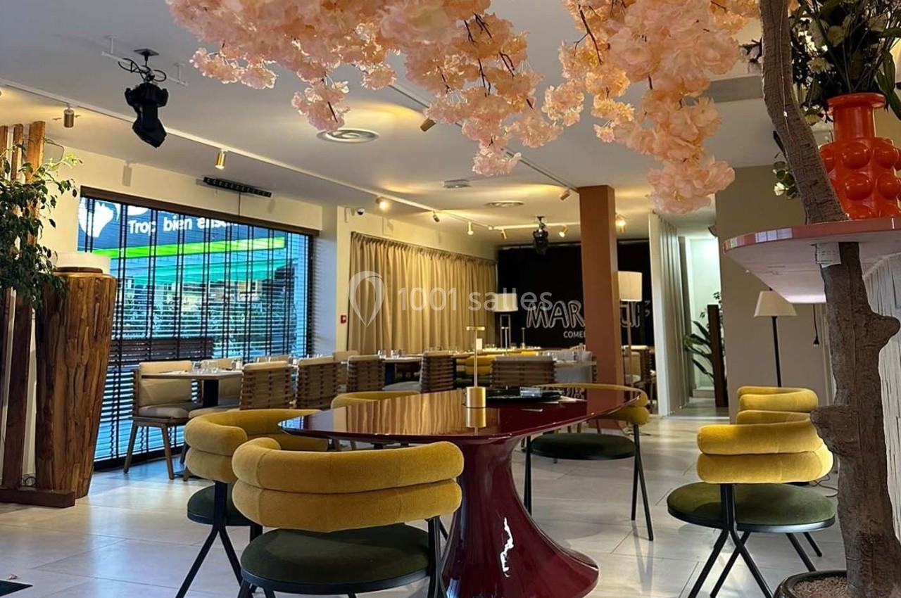 Salle de restaurant moderne avec mobilier coloré, table rouge, chaises jaunes et décoration florale suspendue.