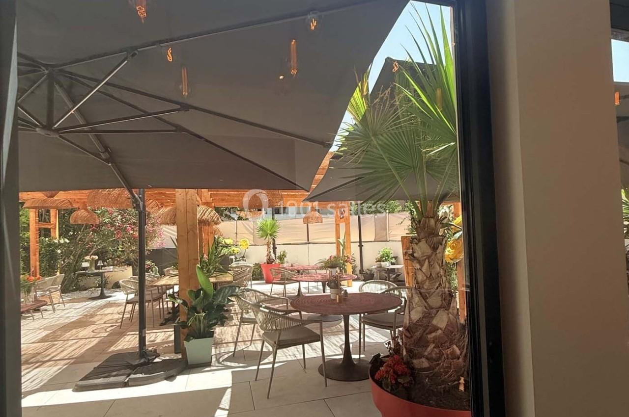 Terrasse extérieure avec tables, chaises, parasols et plantes, éclairée par une lumière naturelle.