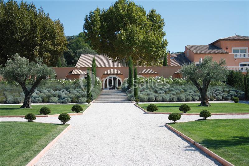 Allée gravillonnée bordée d'arbres et de buissons menant à une maison aux murs ocres entourée de végétation.
