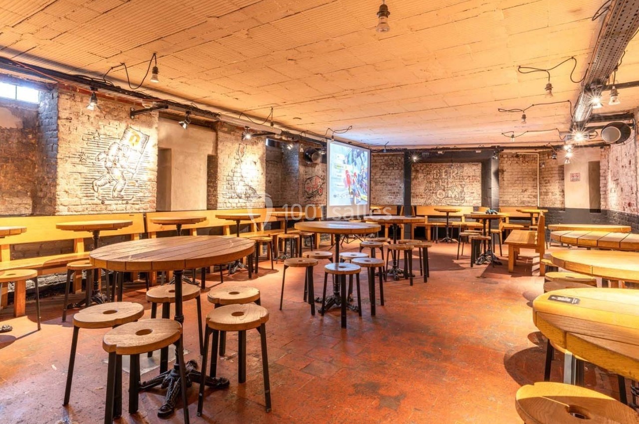Salle avec murs en briques, tables rondes en bois, tabourets, éclairage suspendu et écran de projection au fond.