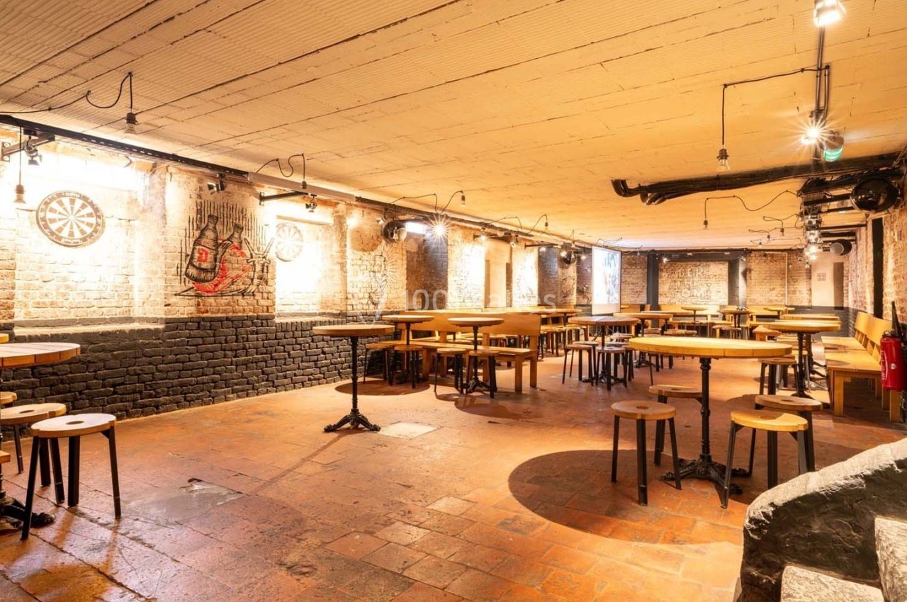 Salle voûtée en briques avec tables et tabourets en bois, éclairée par des lumières suspendues.