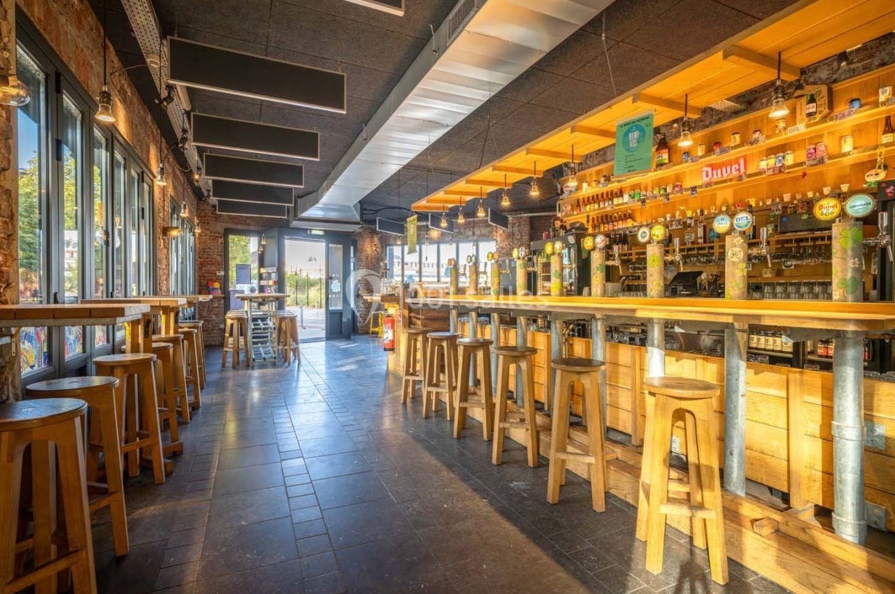 Intérieur d'un bar lumineux avec un long comptoir en bois, des tabourets hauts et des étagères garnies de bouteilles.
