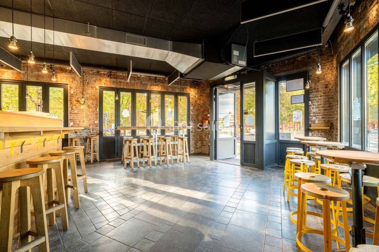 Salle de restaurant lumineuse avec murs en briques, grandes fenêtres, tables et tabourets en bois clair.