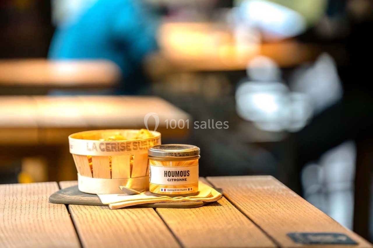 Un pot de houmous citron et des couverts en bois posés sur une table en bois dans un environnement lumineux.