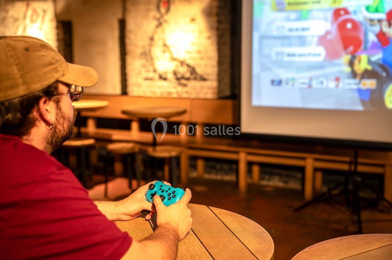 Un homme joue à un jeu vidéo sur un grand écran dans un espace intérieur avec des tables en bois.
