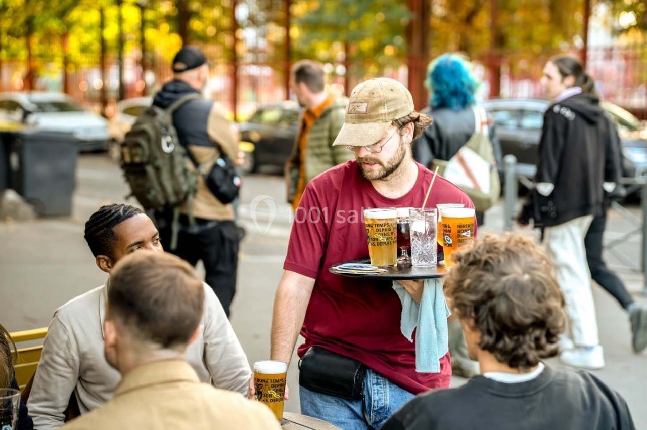 Serveur apportant des verres de bière à une table en terrasse, entourée de clients discutant en plein air.