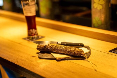 Un couteau et un saucisson sec posé sur une planche en bois, avec un verre de bière en arrière-plan.