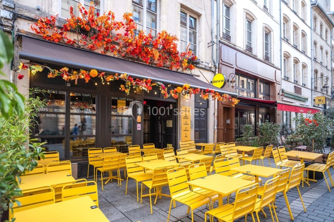 Terrasse d'un café avec des tables et chaises jaunes, décorée de fleurs rouges et oranges, dans une rue piétonne.