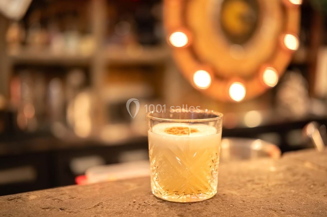 Verre de cocktail avec mousse et tranche de citron séché posé sur un comptoir dans un bar à l'ambiance tamisée.