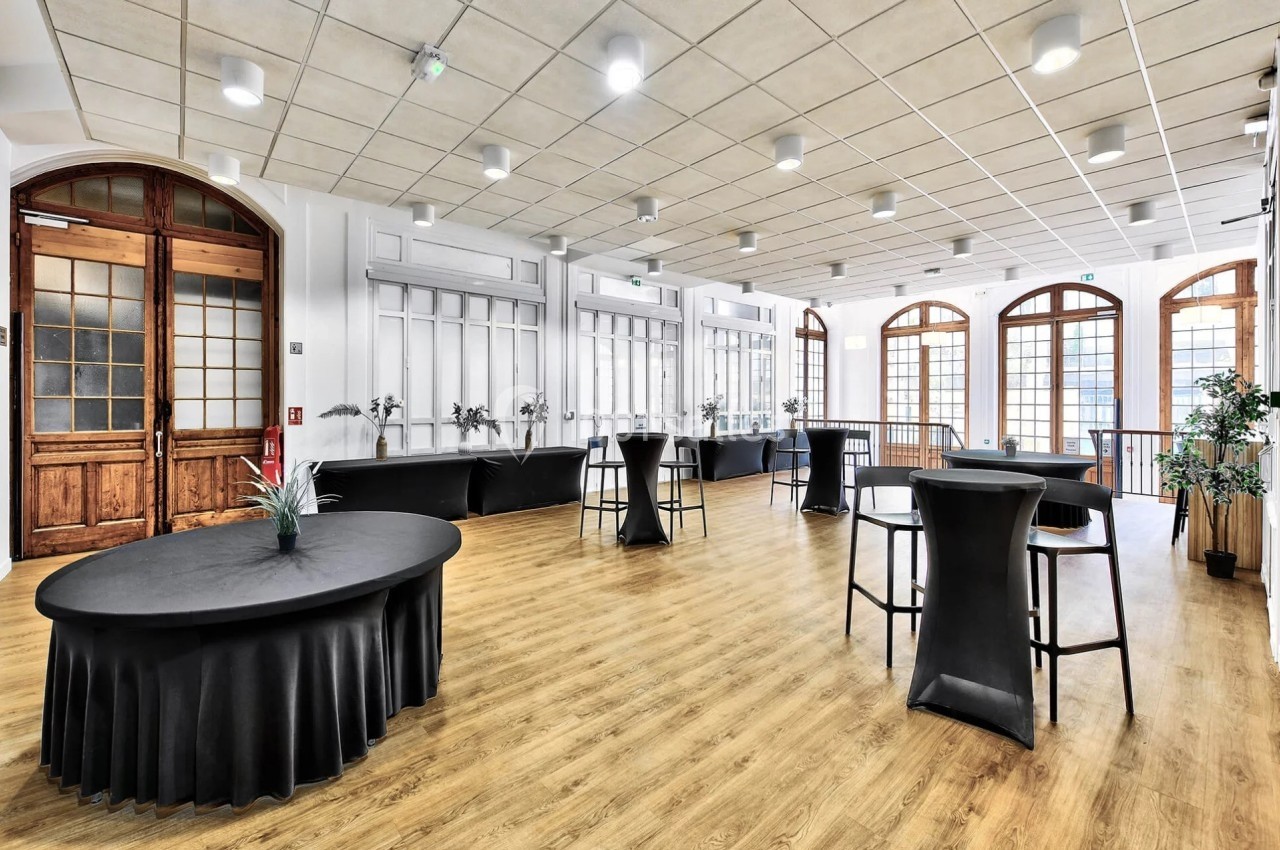 Salle lumineuse avec parquet, grandes fenêtres cintrées, tables rondes et hautes noires, et plantes décoratives.
