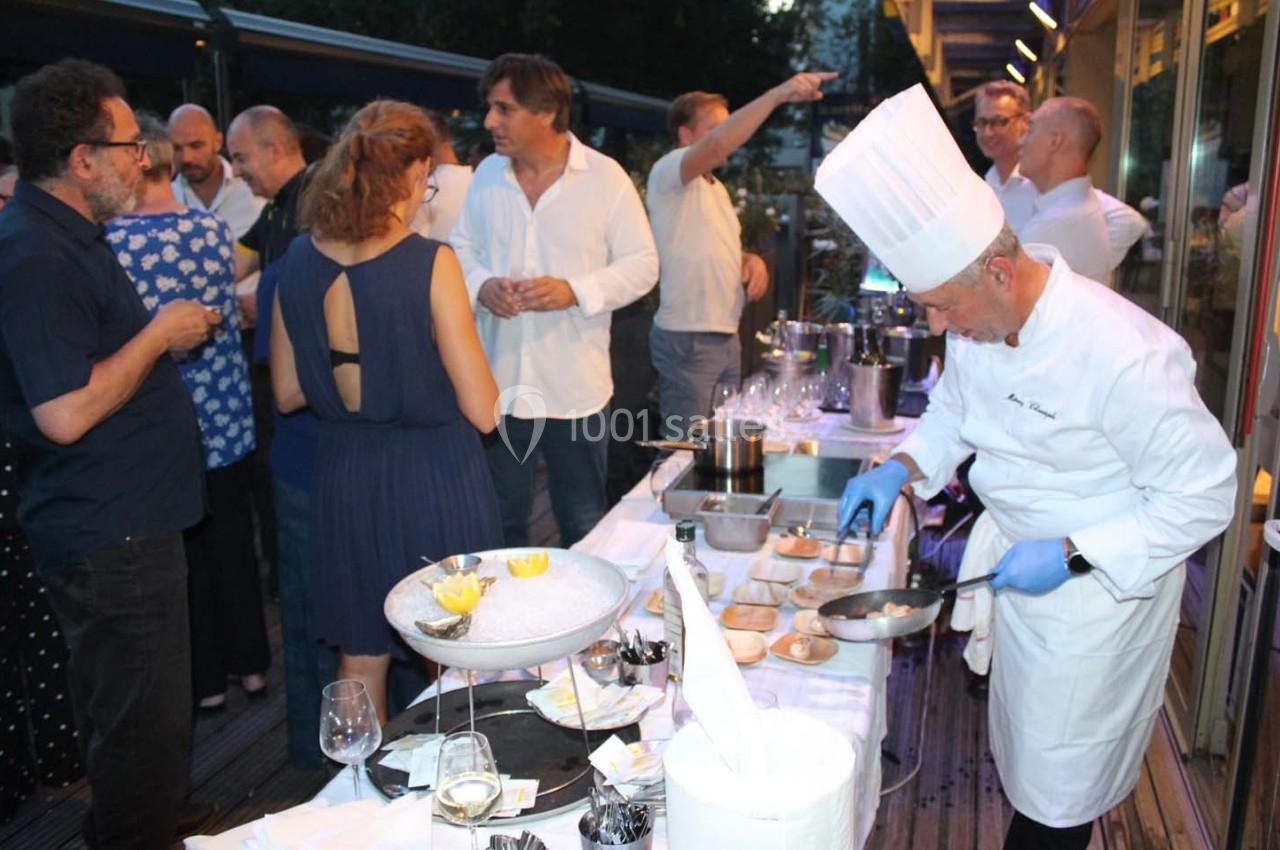 Un chef en tenue blanche prépare des plats lors d'un événement en extérieur, entouré de convives discutant.