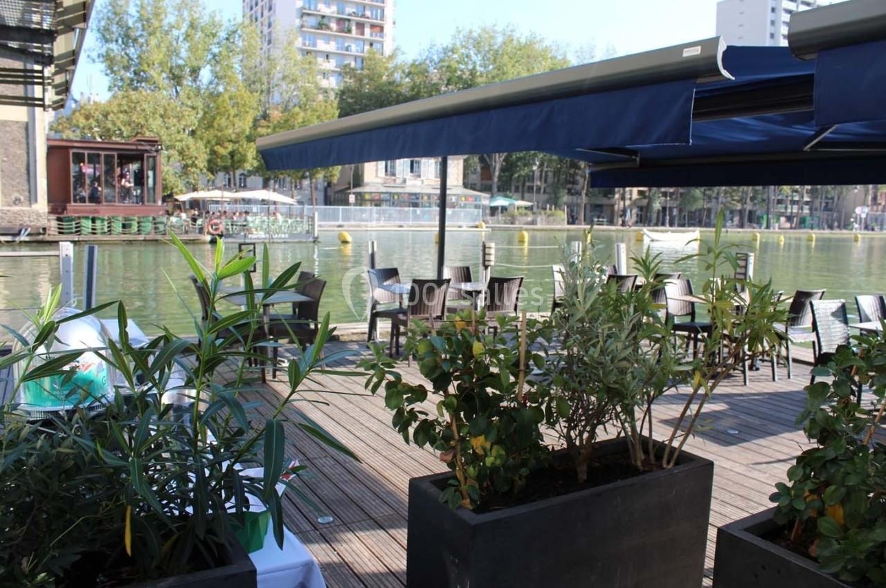 Terrasse en bois avec des plantes en pot, tables et chaises, donnant sur un canal bordé d'arbres et de bâtiments.