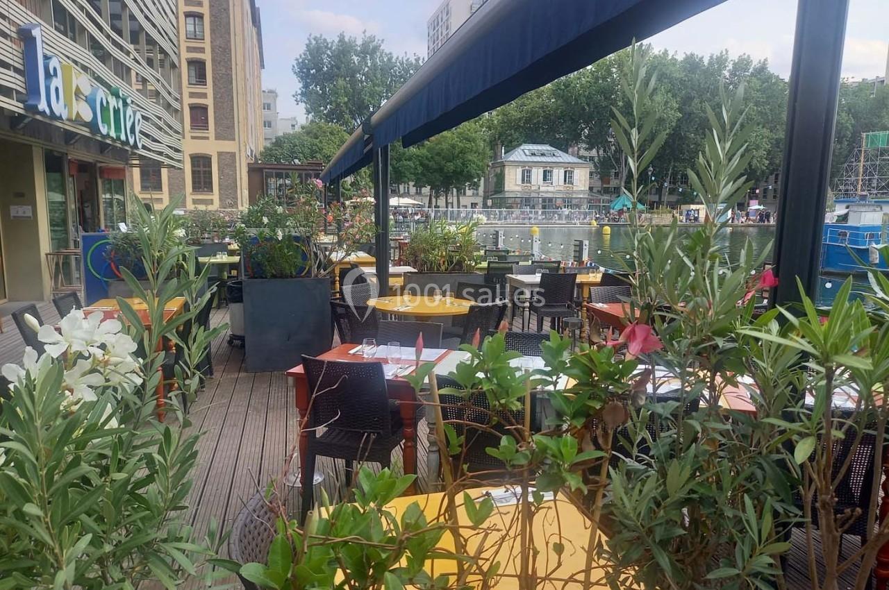 Terrasse d’un restaurant avec tables dressées, plantes en pot et vue sur un canal bordé d’arbres.
