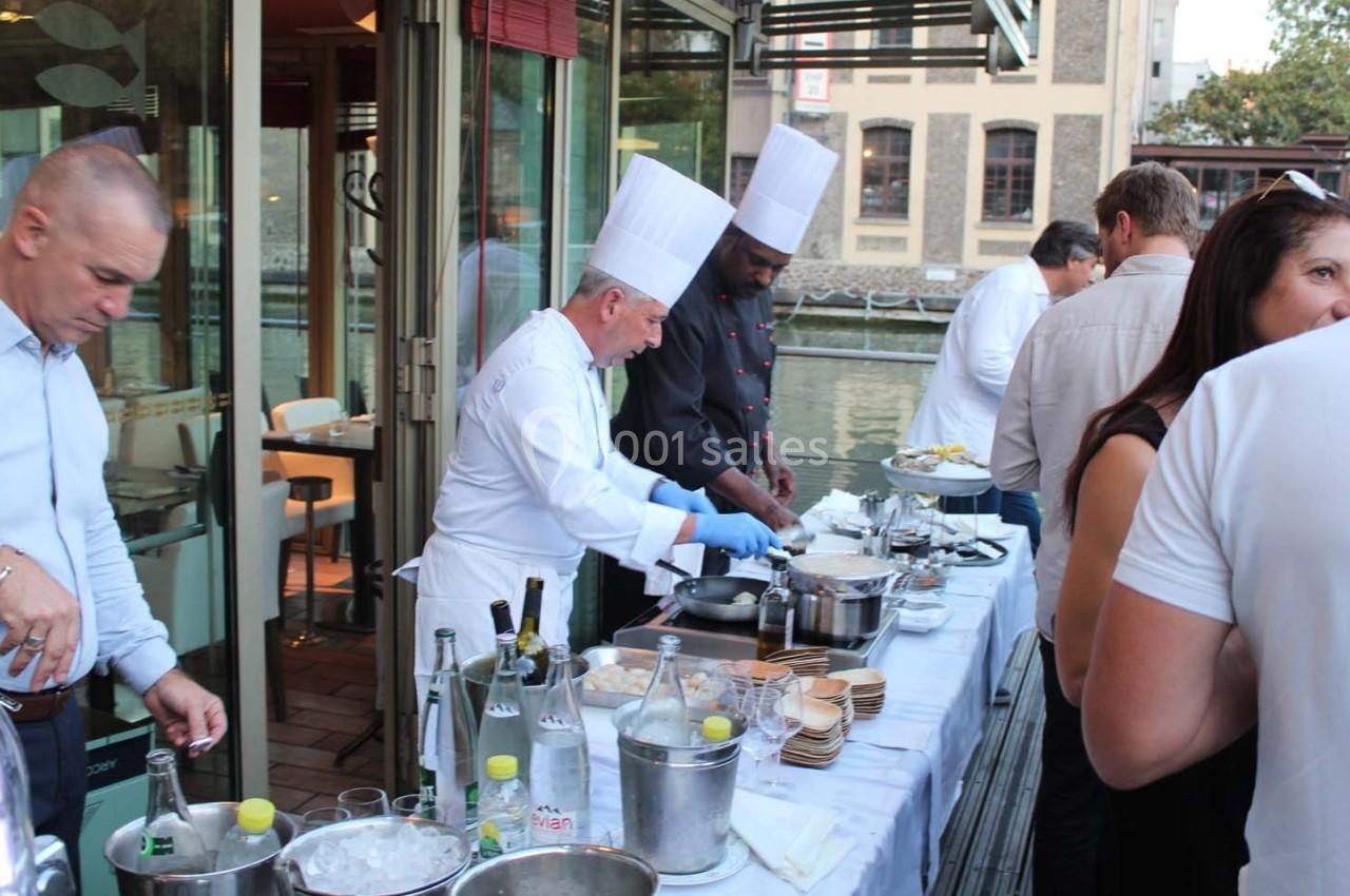 Des chefs cuisinent en extérieur sur une terrasse près d'un canal, entourés de convives et de tables dressées.