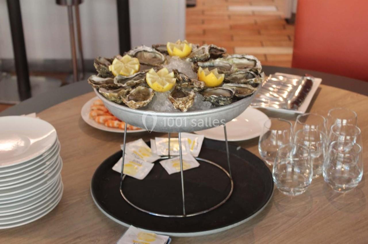 Plateau d'huîtres sur glace avec quartiers de citron, entouré de verres, d'assiettes et de crevettes sur une table.