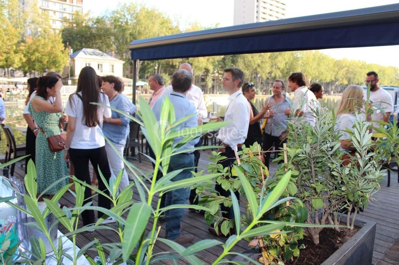 Groupe de personnes discutant sur une terrasse en bois bordée de plantes, avec des arbres et des bâtiments en arrière-plan.